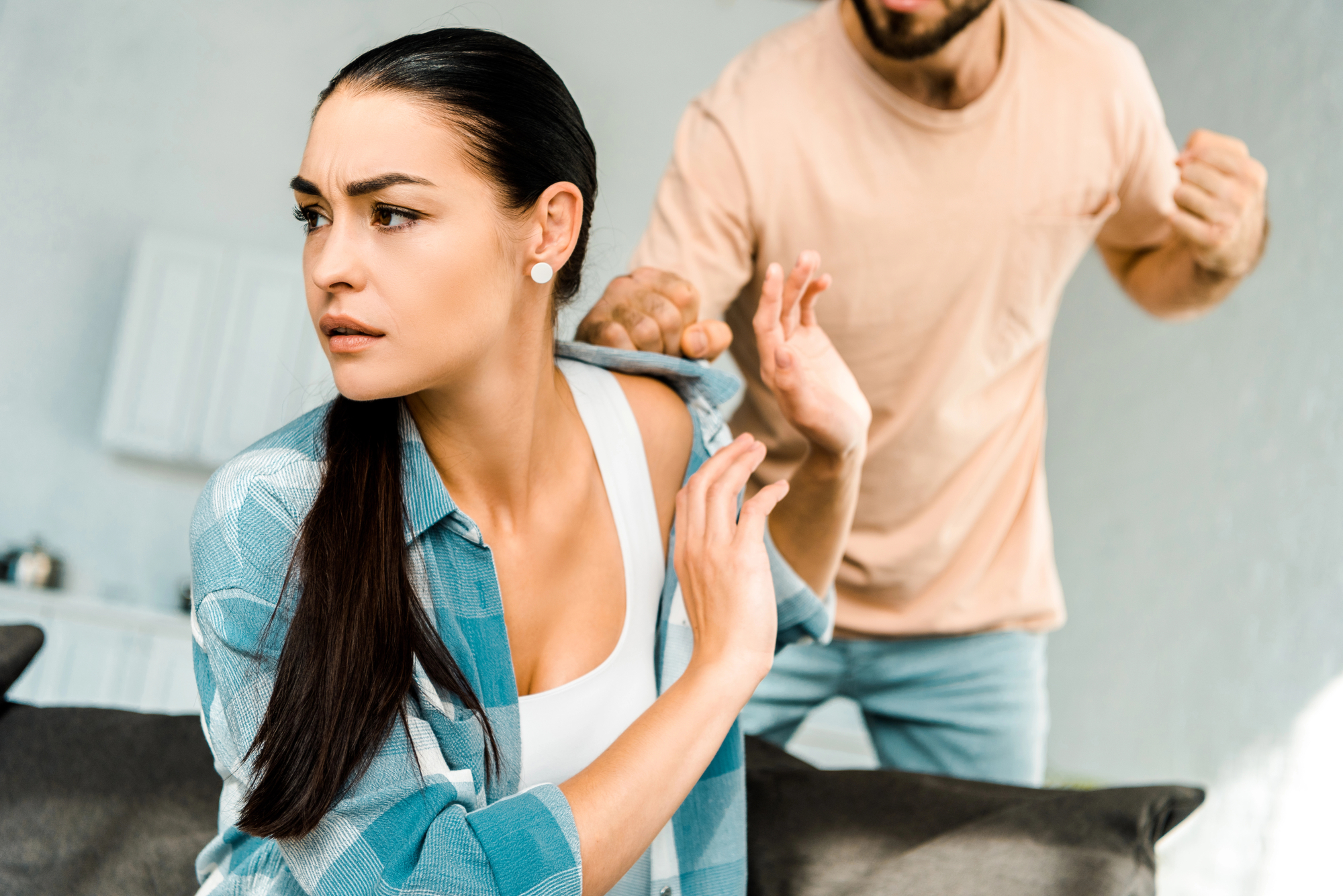 A woman with long dark hair and a concerned expression is turning away and raising her hands defensively as a man in a peach-colored shirt and gray pants stands behind her with his fists clenched. They appear to be in a domestic setting.