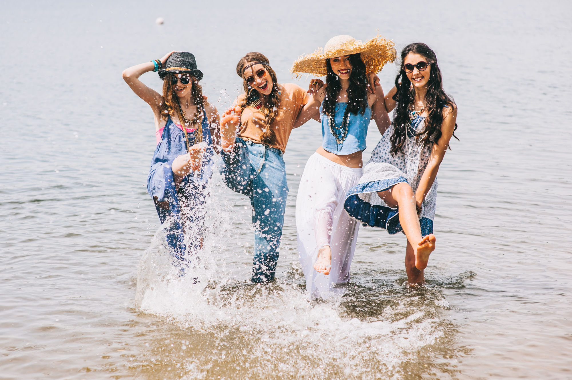 Four women are joyfully splashing water as they stand knee-deep at the beach. They wear trendy summer outfits including hats, overalls, and sunglasses. They all have wide smiles, capturing a moment of fun and camaraderie. The background is a calm body of water.