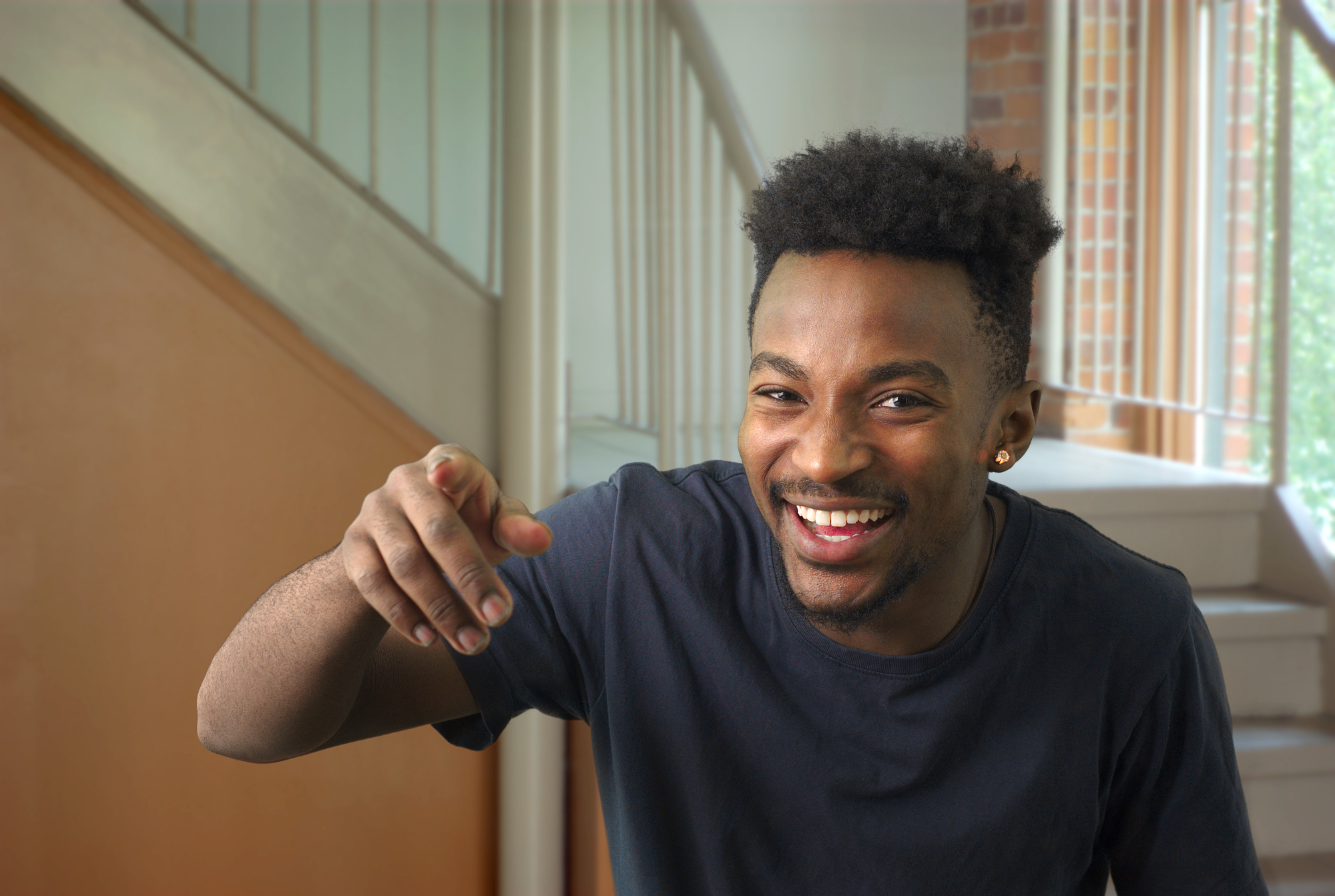 A smiling man with short curly hair and wearing a dark t-shirt is pointing towards the camera. He is standing indoors near a staircase with a brick wall in the background. His expression is joyful and engaging.