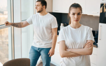 A woman with her arms crossed stands in a modern kitchen, looking directly at the camera with a serious expression. In the background, a man with a beard looks out of a large window, leaning on the windowsill. Both are wearing white t-shirts and jeans.