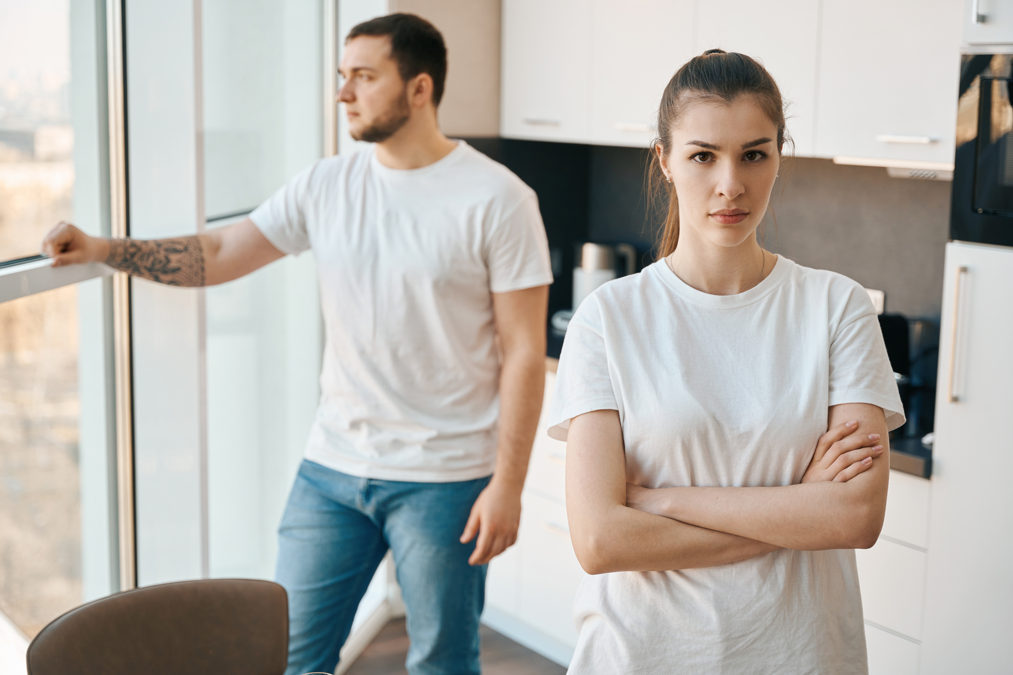 A woman with her arms crossed stands in a modern kitchen, looking directly at the camera with a serious expression. In the background, a man with a beard looks out of a large window, leaning on the windowsill. Both are wearing white t-shirts and jeans.