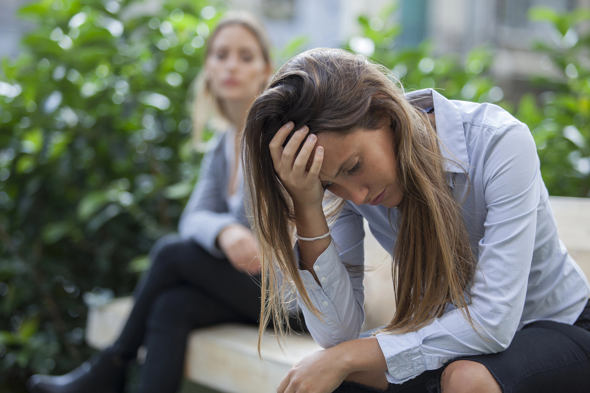 Two women are sitting on a bench outdoors. The woman in the foreground looks upset, resting her head on her hand. The woman in the background is looking at her with a concerned expression. There is green foliage in the background.