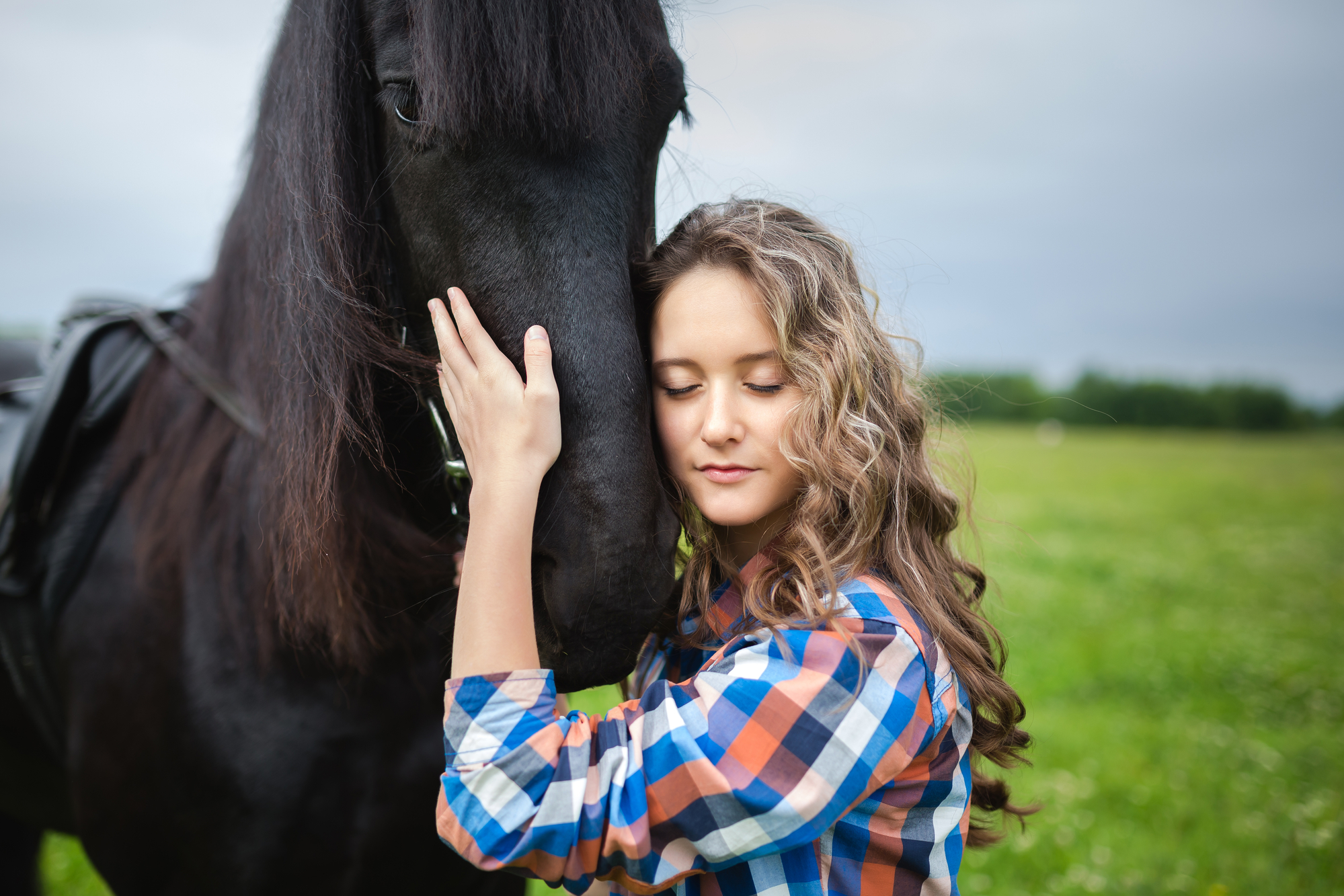 A young woman gently embraces the head of a large black horse while standing in a lush green field. She is wearing a plaid shirt, and her long curly hair cascades over her shoulder. The sky is overcast, creating a calm and serene atmosphere.