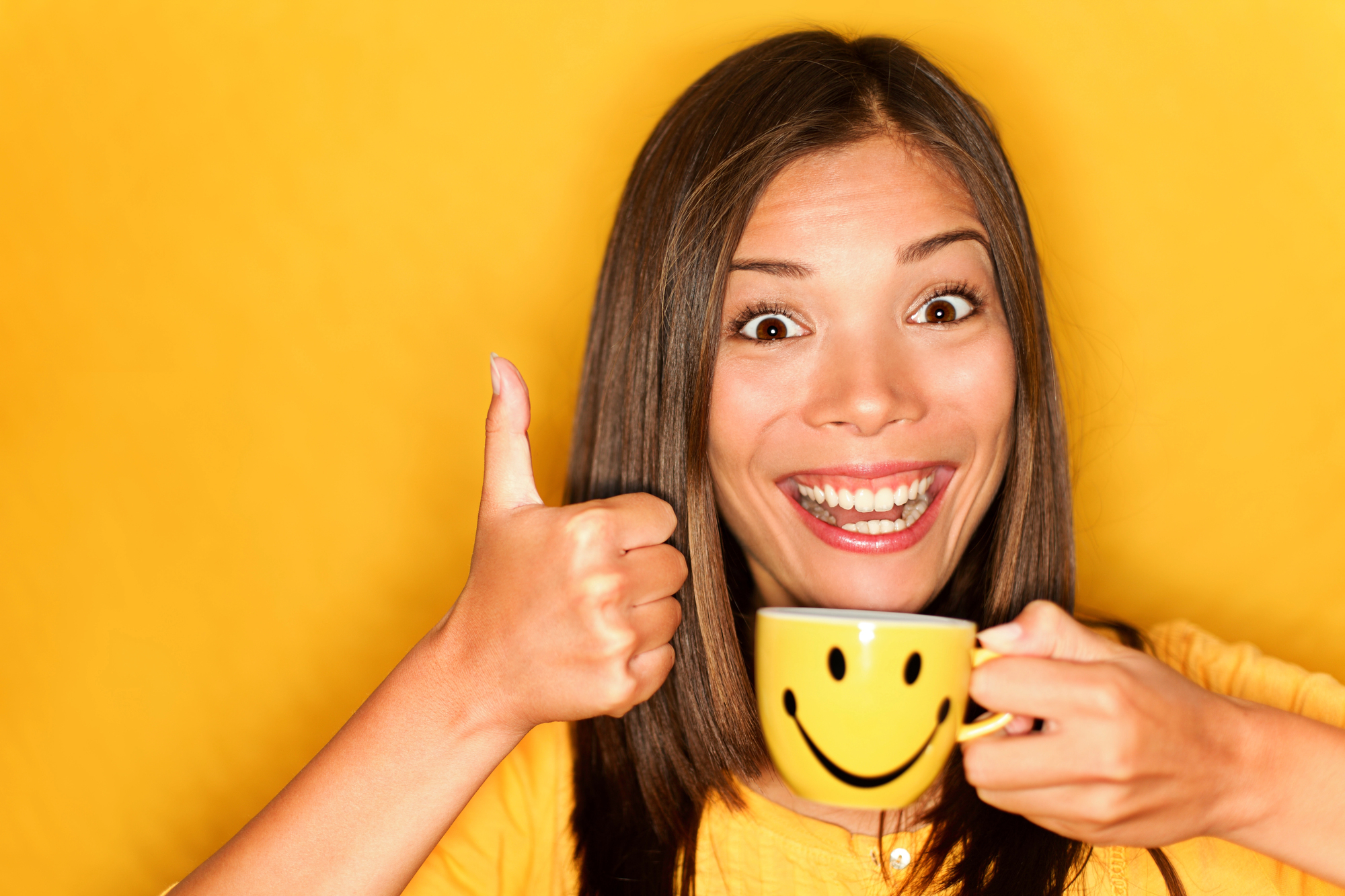 A woman with long brown hair smiles widely while holding a yellow mug with a smiley face on it. She is wearing a yellow shirt and giving a thumbs-up gesture with her other hand. The background is yellow, matching her shirt and the mug.