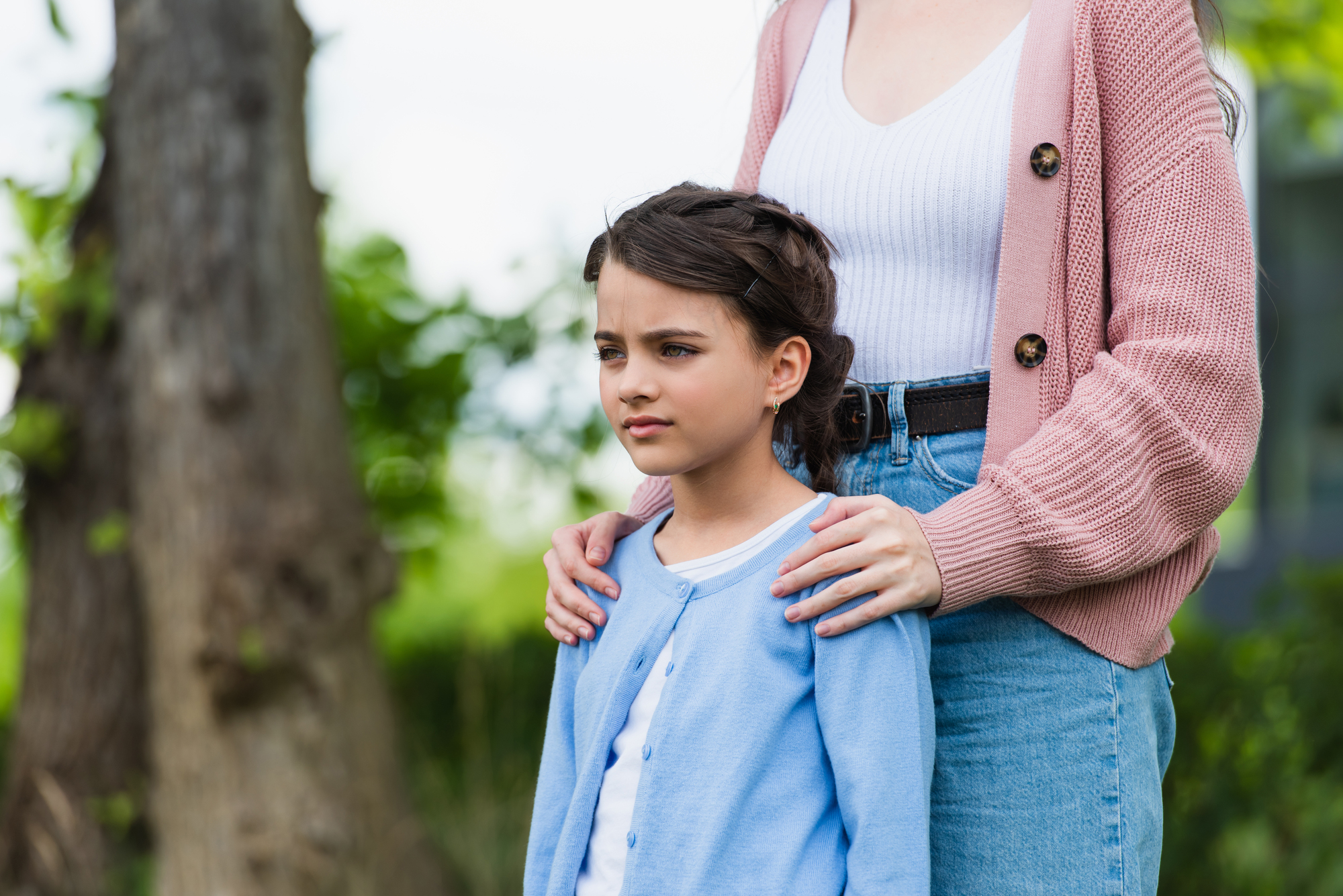 A young girl in a light blue cardigan and white shirt stands in front of an adult woman in a pink cardigan and white top, who has her hands gently resting on the girl's shoulders. They are outside, with green foliage and trees blurred in the background.