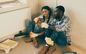 A couple takes a break while renovating, sitting on the floor by a wall. Both are wearing casual clothes and work gloves. The man is offering the woman a drink, and they both look happy and relaxed. Renovation tools and paint supplies are scattered around them.