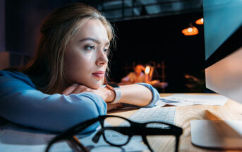 A young woman with long blonde hair is resting her chin on her hands, gazing intently at a computer screen in a dimly lit room. She has a serious expression. In the foreground, a pair of eyeglasses are placed on the wooden desk beside a keyboard.