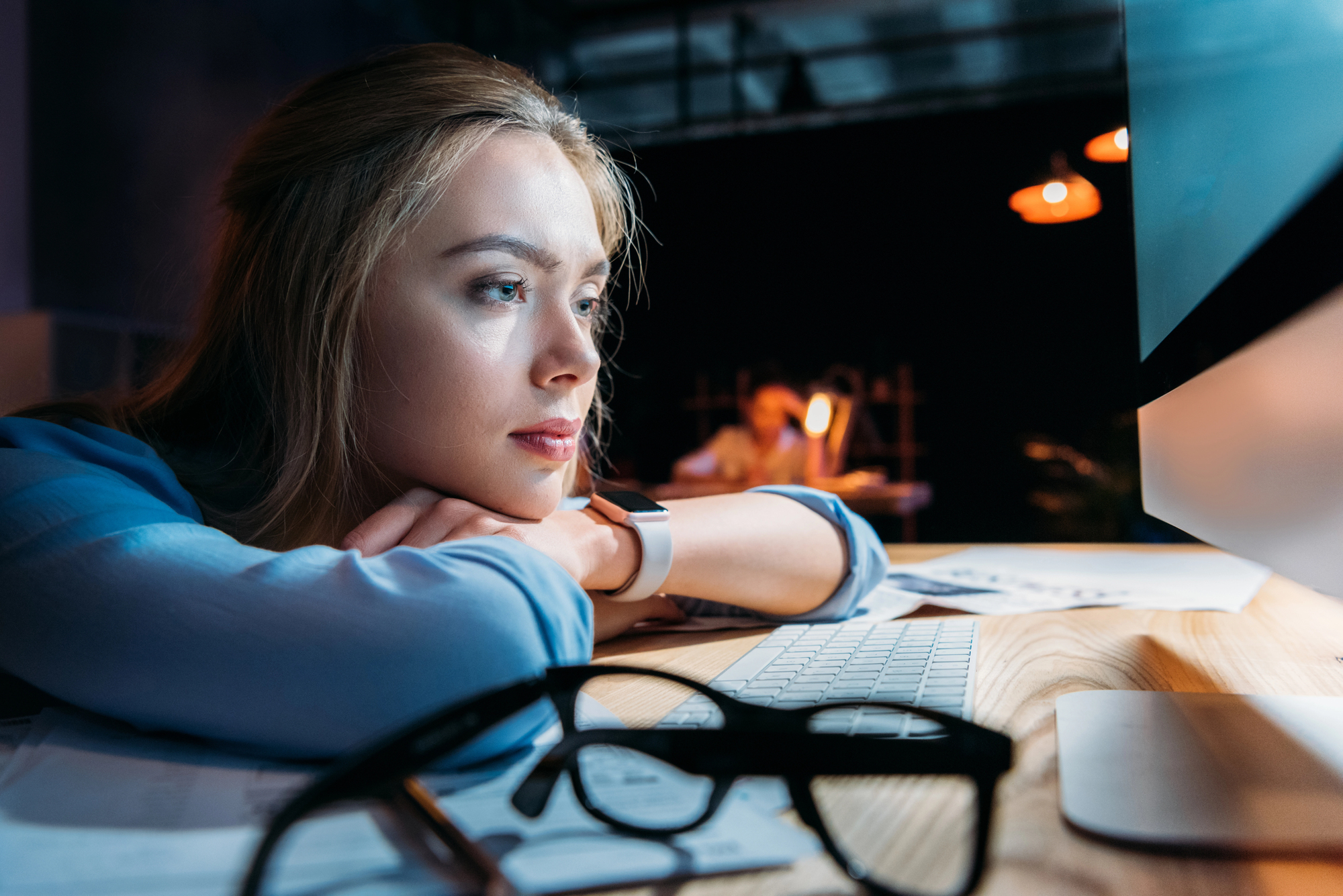 A young woman with long blonde hair is resting her chin on her hands, gazing intently at a computer screen in a dimly lit room. She has a serious expression. In the foreground, a pair of eyeglasses are placed on the wooden desk beside a keyboard.