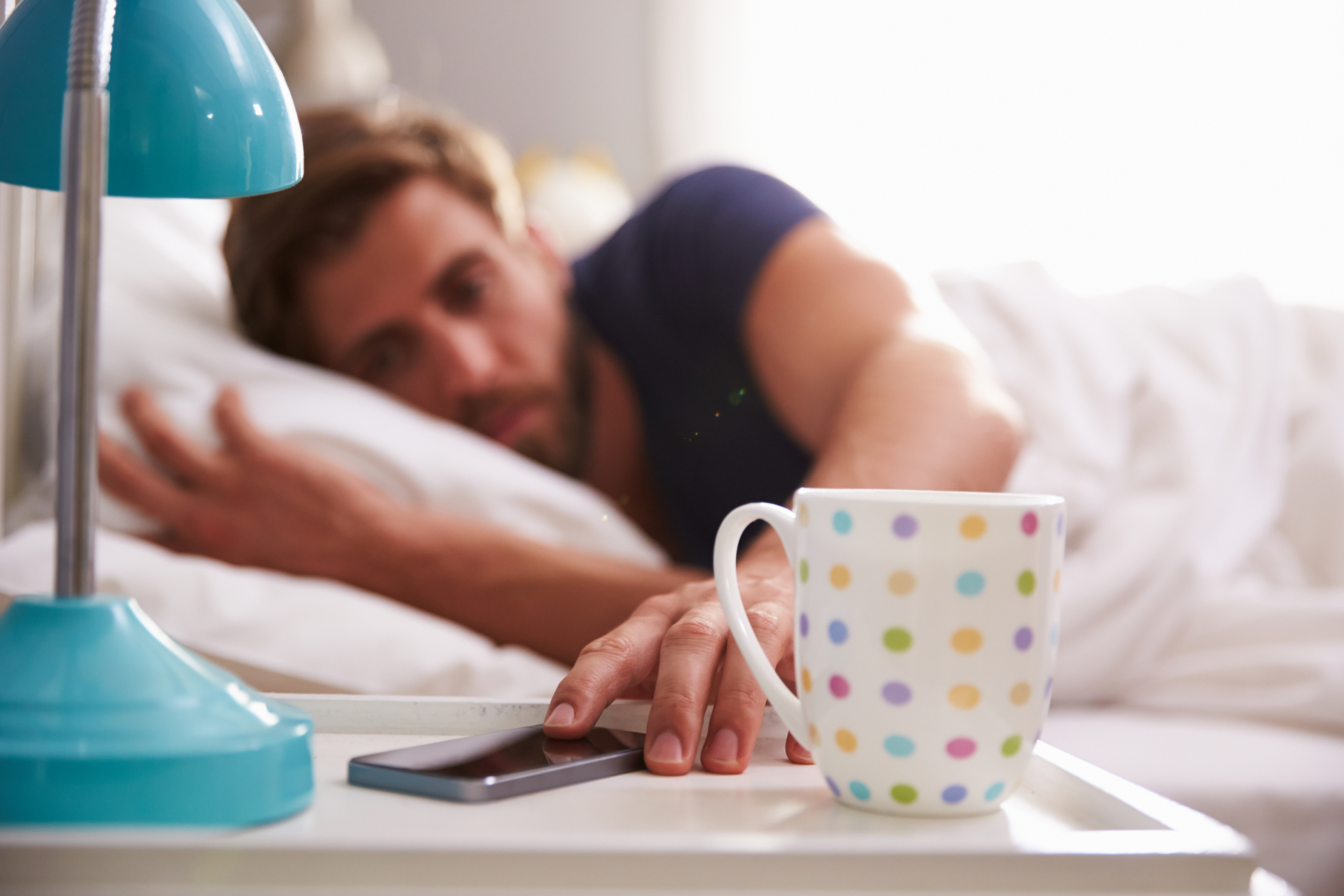 A person lying in bed is reaching for a smartphone on a white bedside table. The table also has a colorful polka dot mug and a blue bedside lamp. The person's facial features are out of focus and they are partially covered by a white blanket.