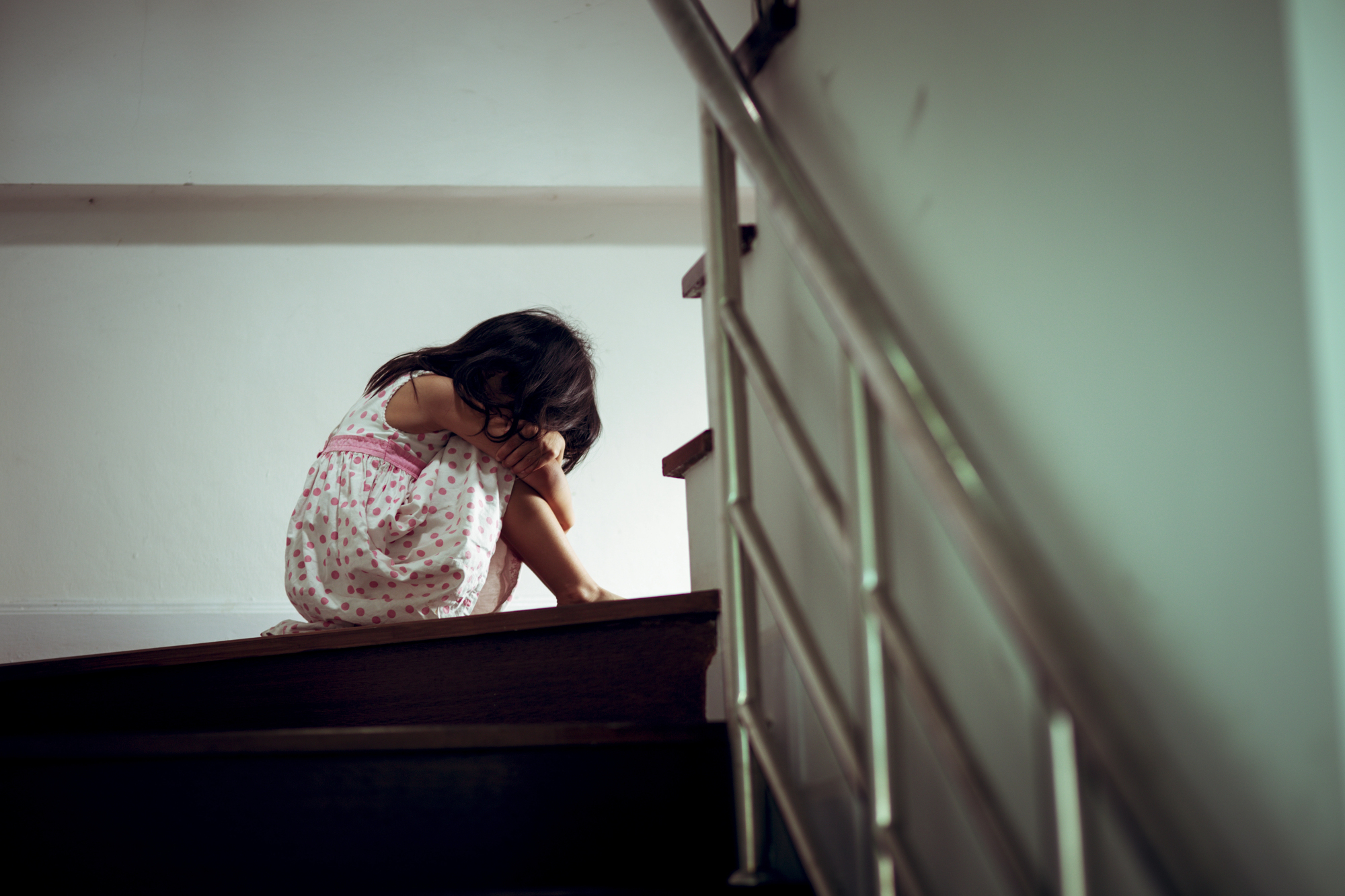 A young girl in a polka-dotted dress sits alone on a staircase with her arms wrapped around her knees and head bowed, creating a sense of sadness or contemplation. The staircase is dimly lit and has a metal handrail.