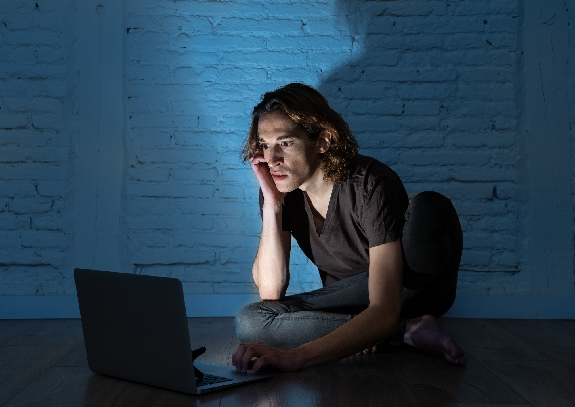 A person with shoulder-length hair sits on the floor in a dimly lit room, leaning forward and staring intently at a laptop. They rest their head on one hand and appear focused, illuminated by the screen's light against a brick wall background.