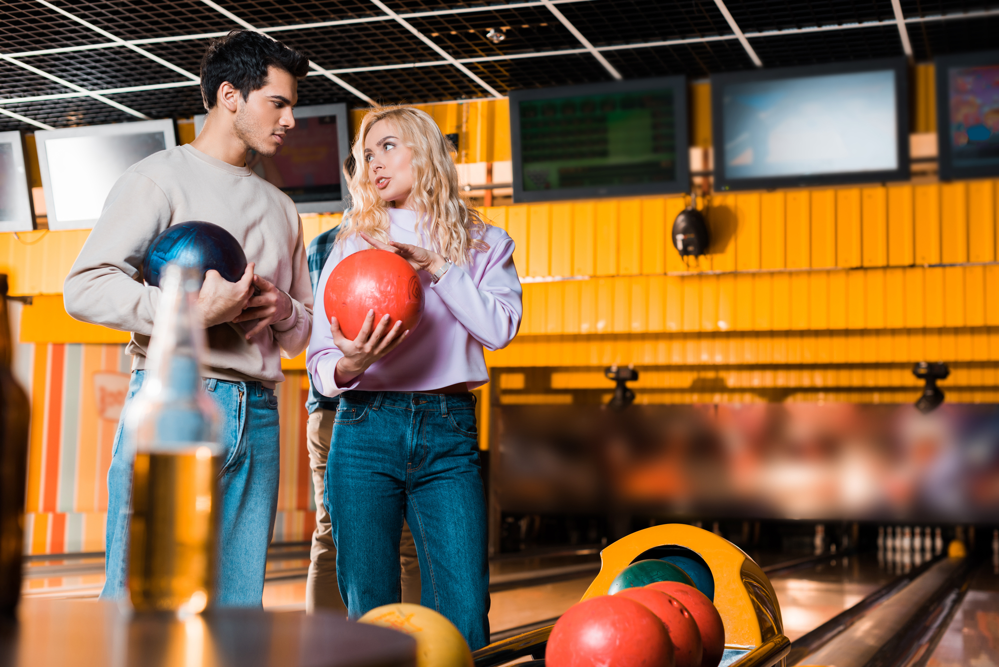 A man and a woman are standing in a bowling alley, each holding a bowling ball. The man is holding a black ball, and the woman is holding a red one. They appear to be conversing. Bowling pins and balls are visible in the foreground, and a beer bottle is on the table.