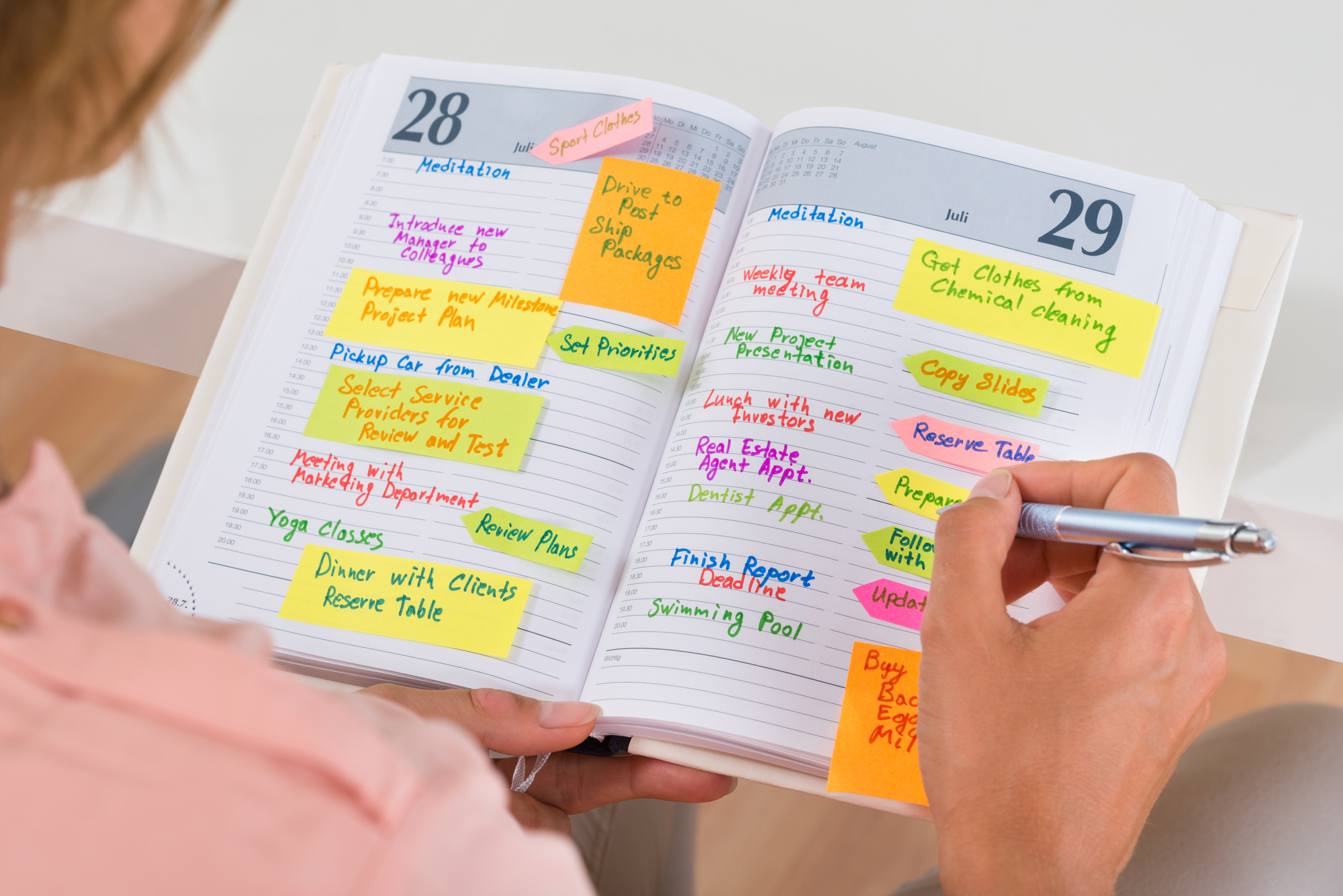 A person holding a pen is looking at an open planner filled with colorful sticky notes and handwritten entries for the 28th and 29th of a month. The notes list various tasks and activities like meetings, yoga classes, and project deadlines.