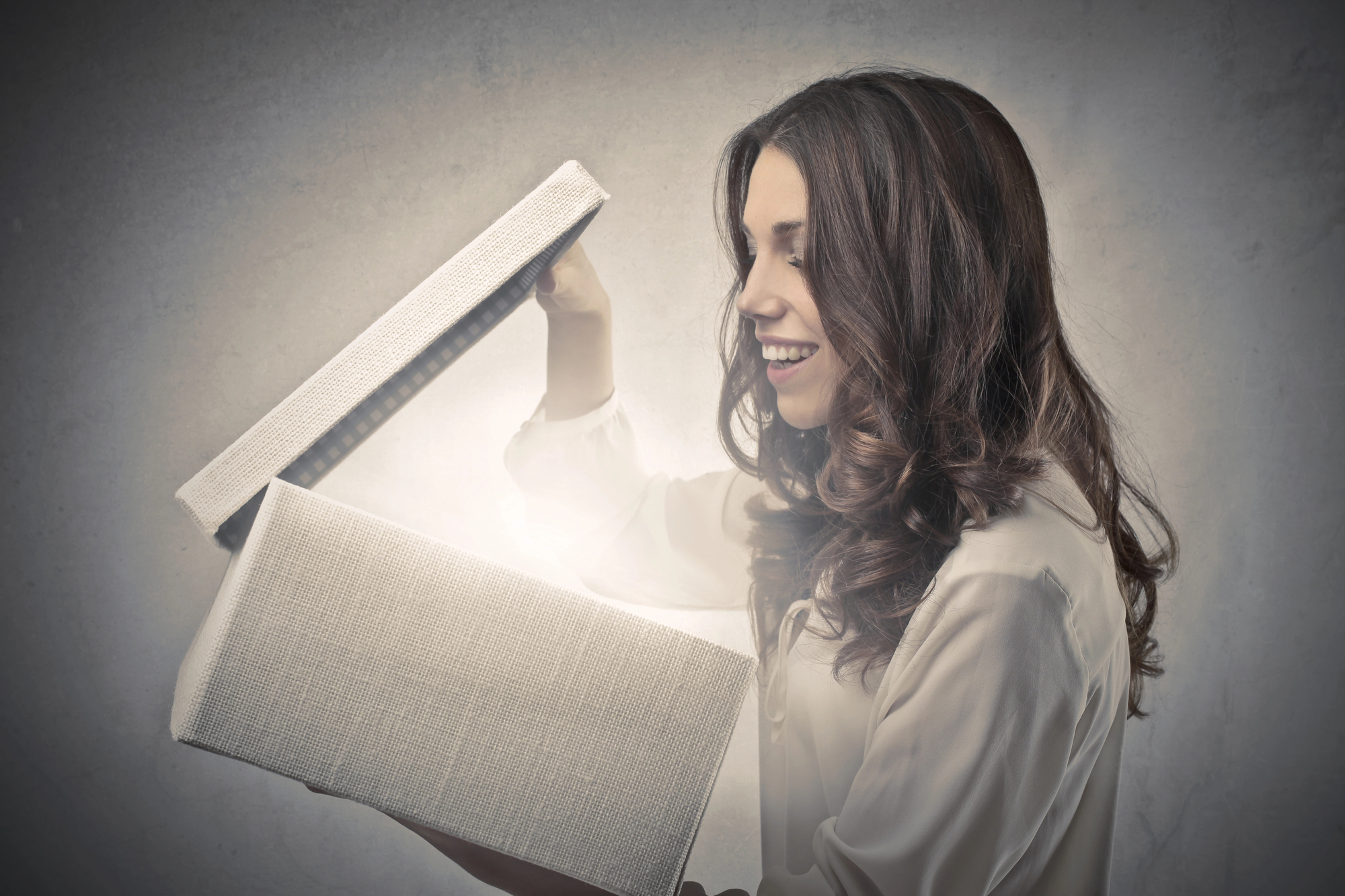 A woman with long, curly hair wearing a white blouse smiles in amazement while opening a large, illuminated gift box. The background is gray and textured, creating a dramatic contrast with the glow from the box.