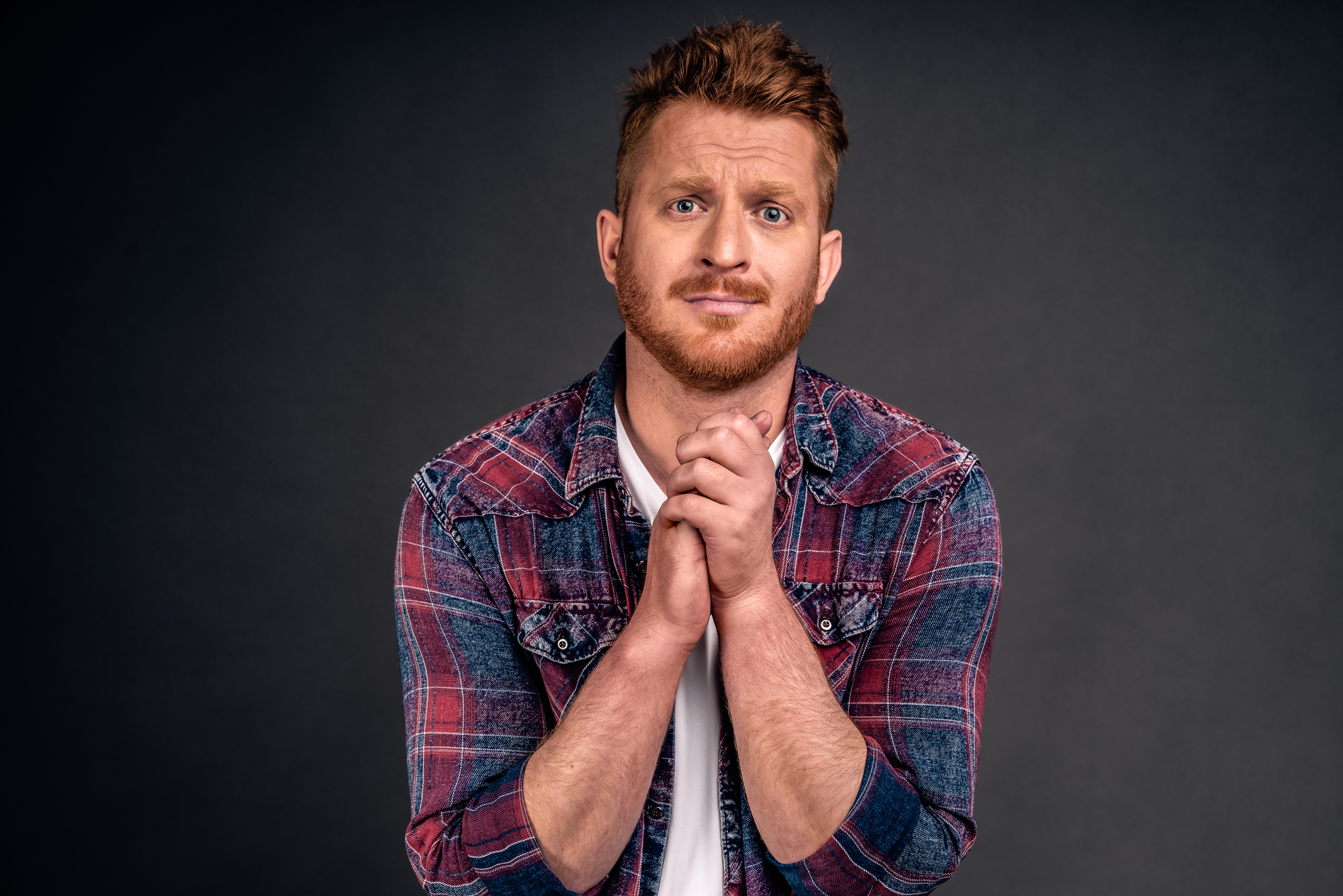 A man with short, reddish hair and a beard is standing against a dark background. He is wearing a red and blue plaid shirt over a white t-shirt. The man has a concerned or worried expression, with his hands clasped together in front of him.