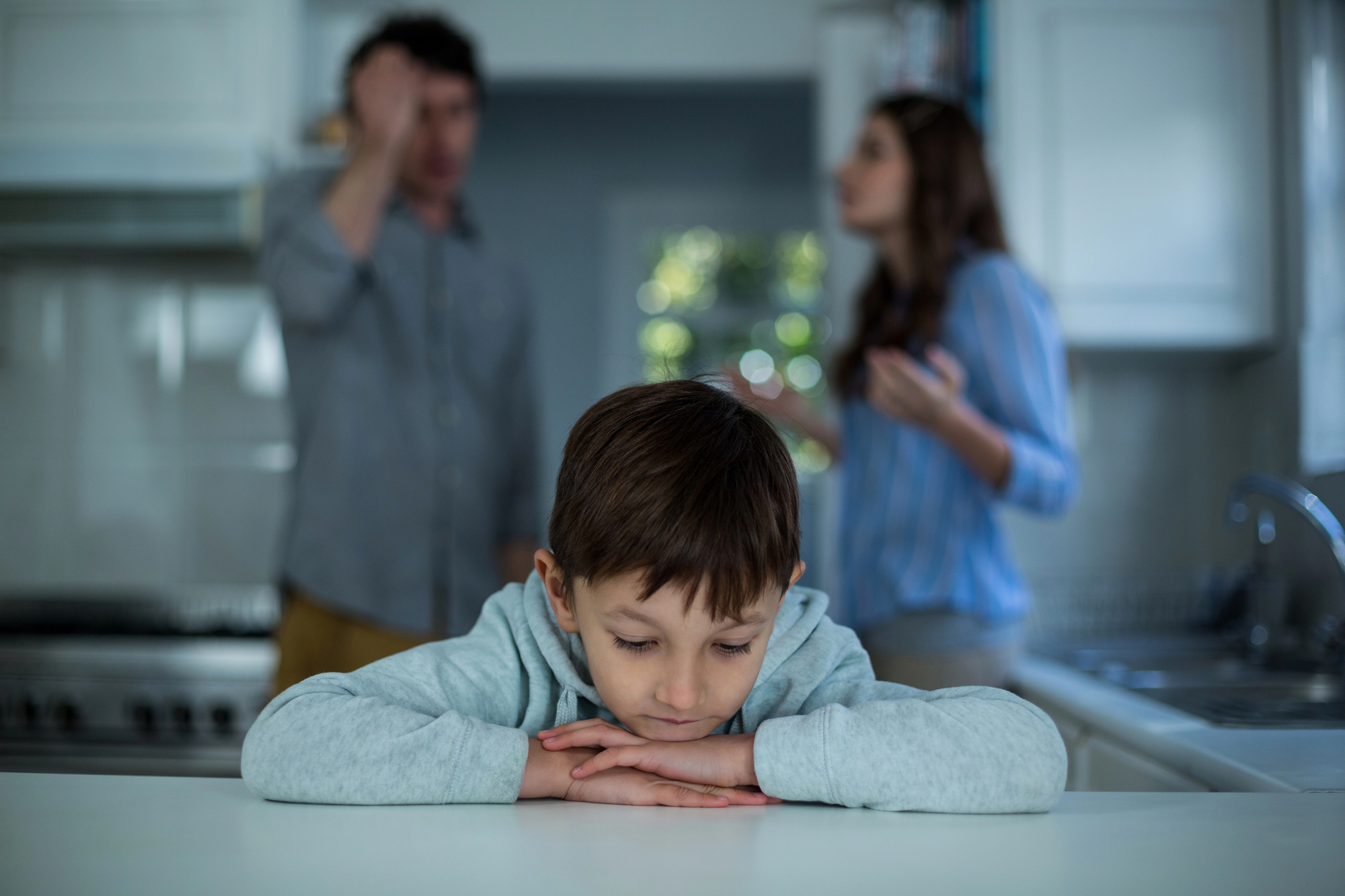 A young boy, looking sad, rests his head on his arms on a kitchen counter while a man and a woman argue in the background. The scene conveys tension and distress in their home environment.