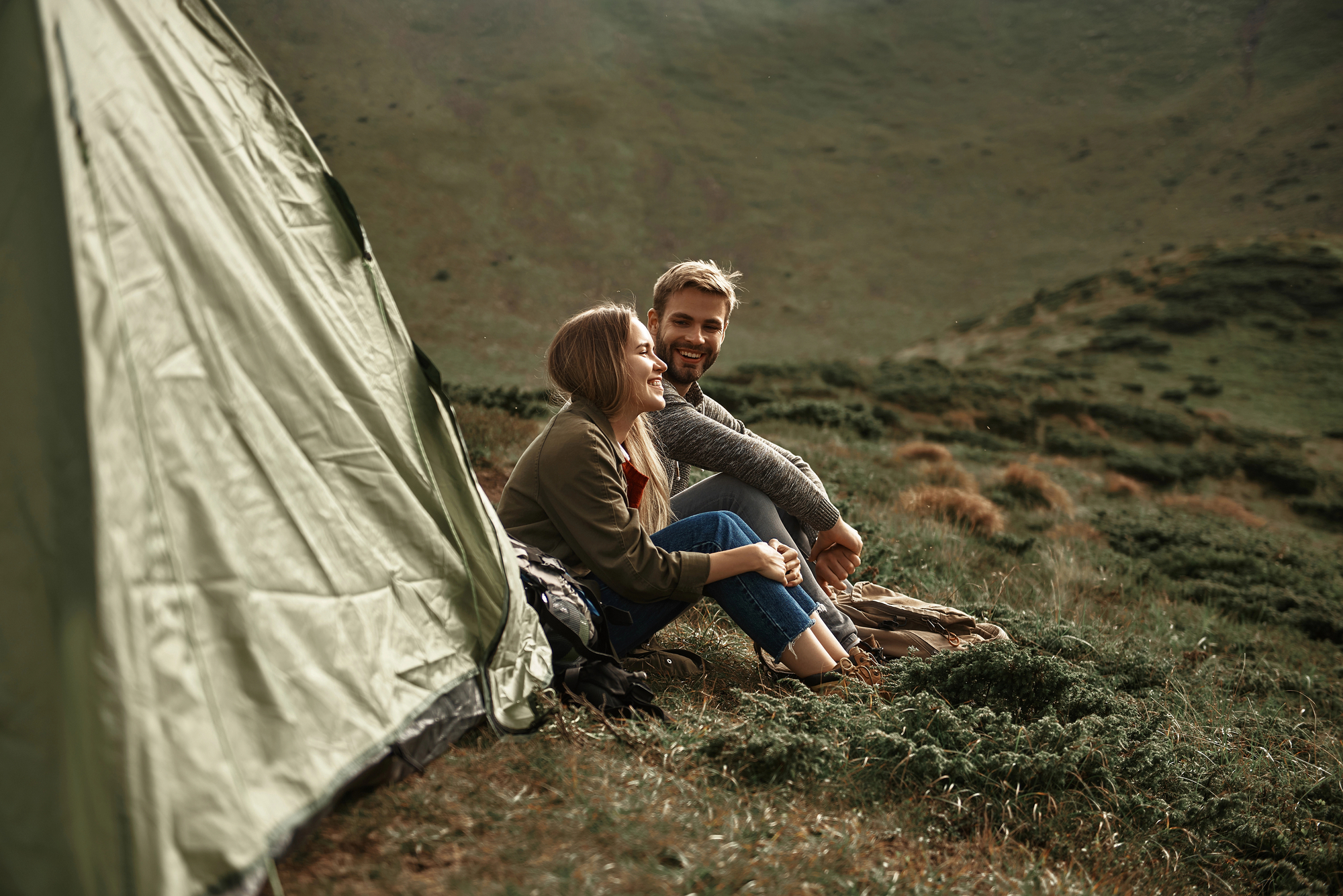 A woman and a man sit beside a green tent in a hilly landscape. Both are smiling and appear to be enjoying their time outdoors. The surroundings are lush with green grass and scattered bushes. The sky is clear, and the atmosphere seems peaceful and relaxed.