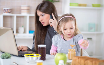 A woman multitasks, talking on the phone and working on a laptop, while a young girl beside her applies peanut butter to a slice of bread. The scene is set in a kitchen with jars of jam, a green apple, and other kitchen items on the counter.