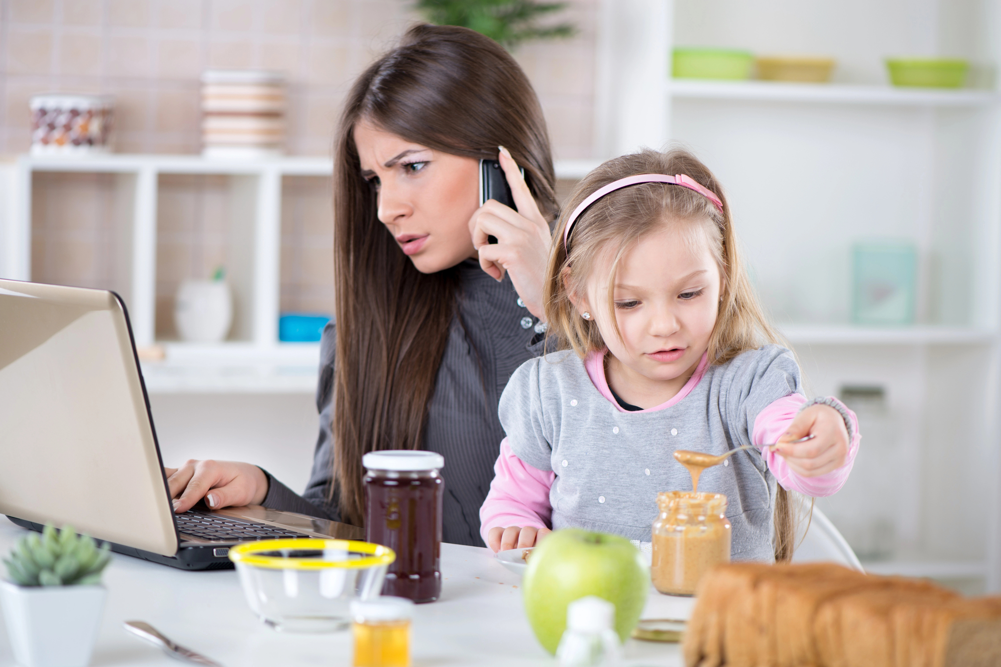 A woman multitasks, talking on the phone and working on a laptop, while a young girl beside her applies peanut butter to a slice of bread. The scene is set in a kitchen with jars of jam, a green apple, and other kitchen items on the counter.