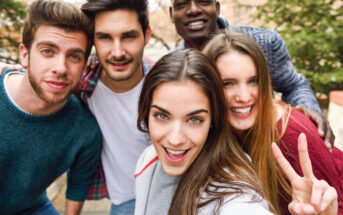 Five friends take a cheerful selfie outdoors. They all smile warmly at the camera, and one person is making a peace sign with their fingers. The group seems to be enjoying a casual day out, with greenery and buildings visible in the background.