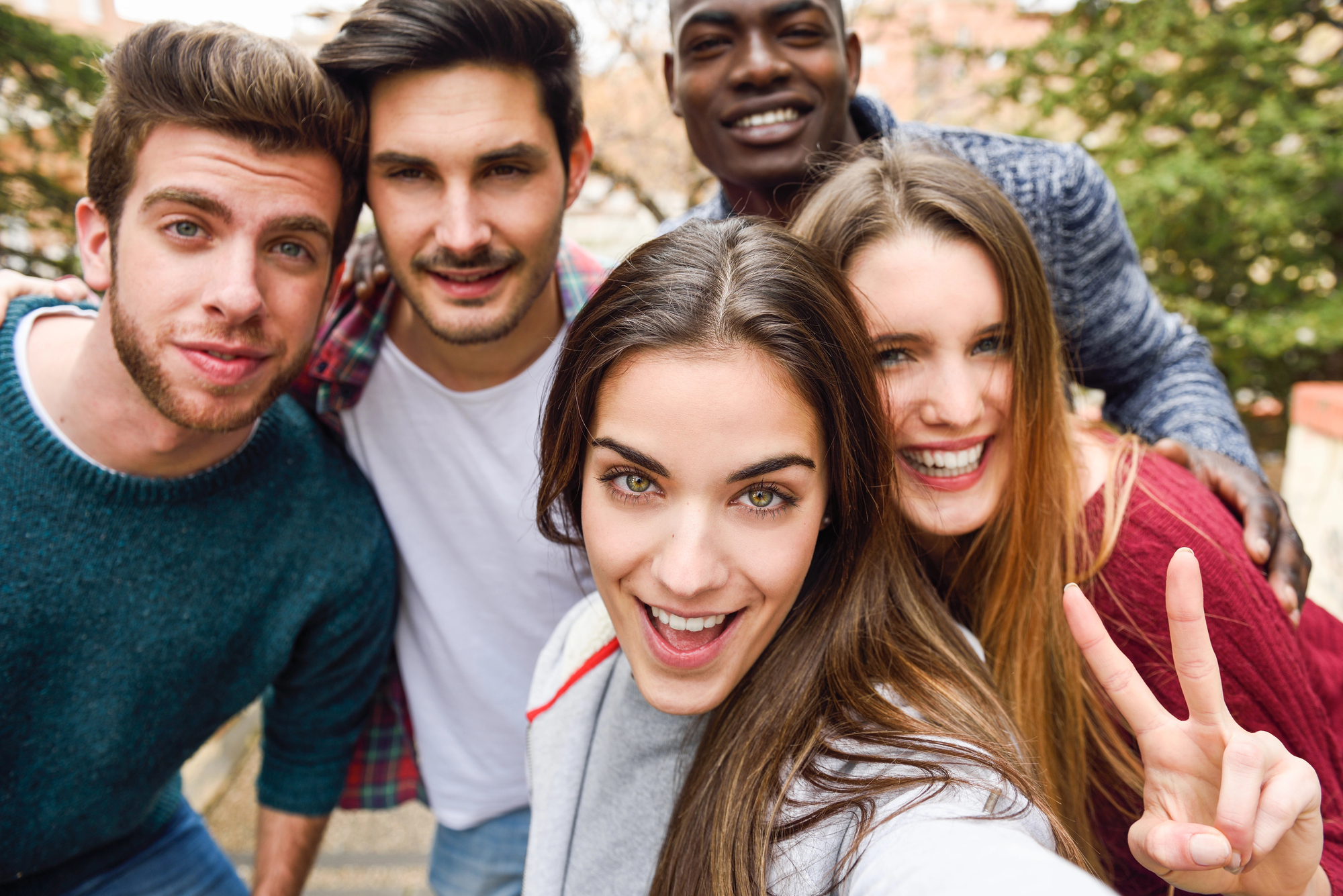 Five friends take a cheerful selfie outdoors. They all smile warmly at the camera, and one person is making a peace sign with their fingers. The group seems to be enjoying a casual day out, with greenery and buildings visible in the background.