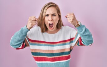 A woman with shoulder-length blonde hair is standing against a light pink background. She is wearing a colorful striped sweater and a watch on her wrist. She looks angry, with her mouth open and fists raised.