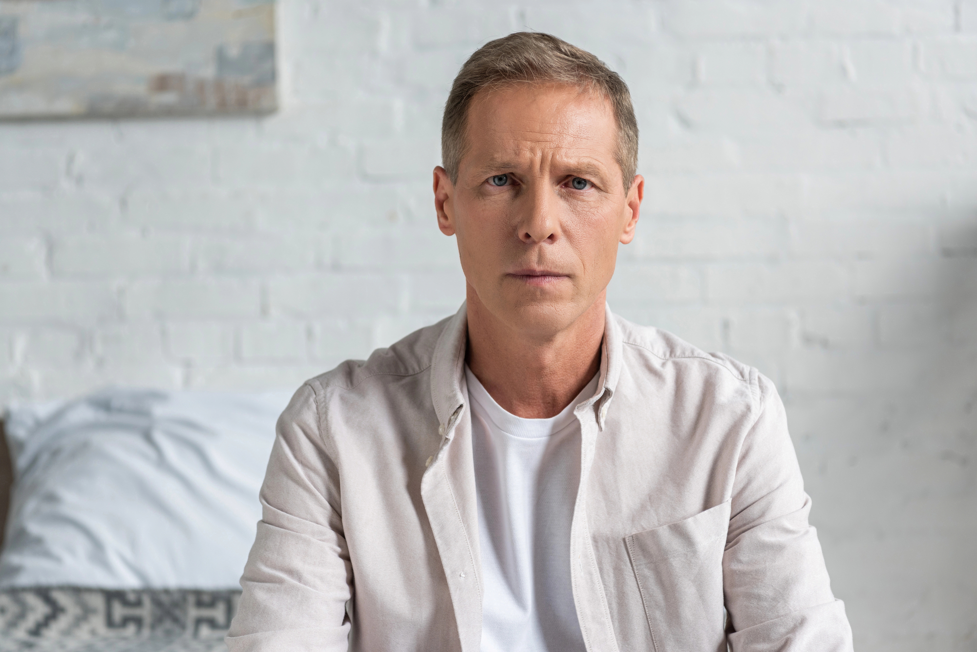 A man with short blond hair and a serious expression is seated in a brightly lit room. He is wearing a white shirt layered with a light beige jacket. A white bed with patterned pillows is visible in the background against a white brick wall.