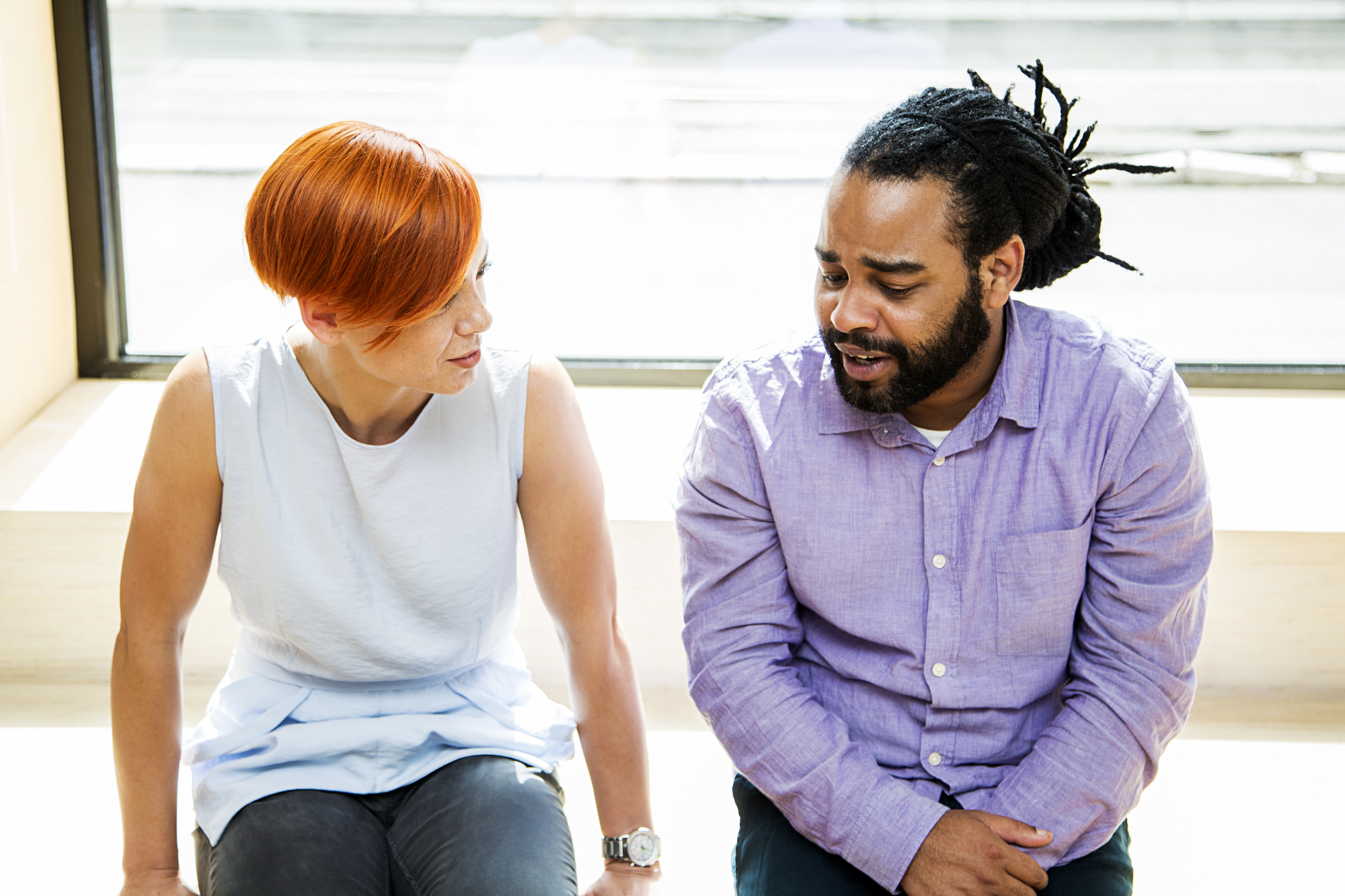 A woman with short red hair wearing a white sleeveless top and a man with dreadlocks wearing a lavender shirt are sitting by a window and engaged in a conversation.