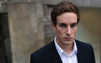 A young man with short brown hair is wearing a white shirt and a dark blazer. He looks directly at the camera with a serious expression. The background is blurred, featuring stone walls and concrete architecture.