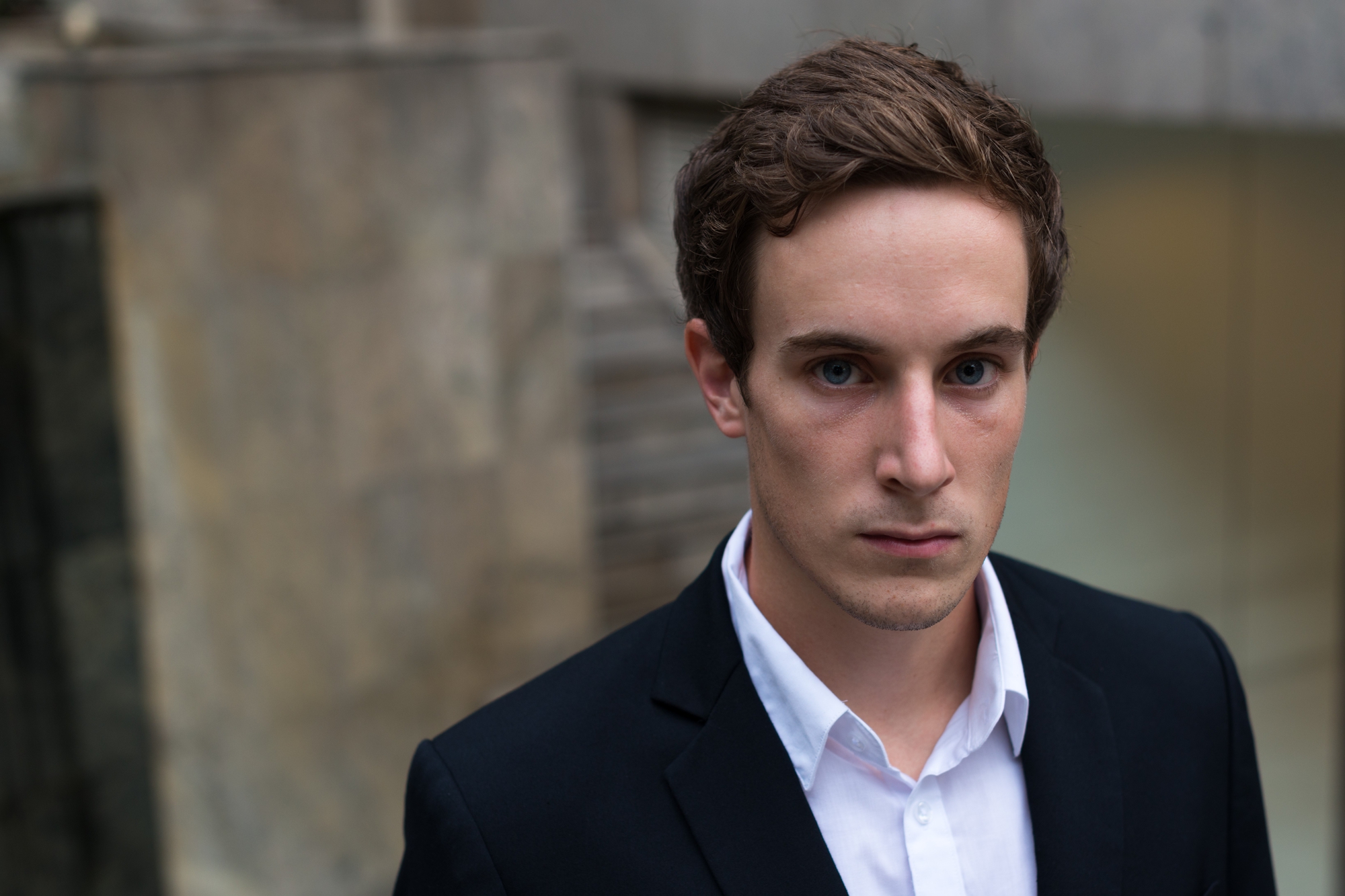 A young man with short brown hair is wearing a white shirt and a dark blazer. He looks directly at the camera with a serious expression. The background is blurred, featuring stone walls and concrete architecture.