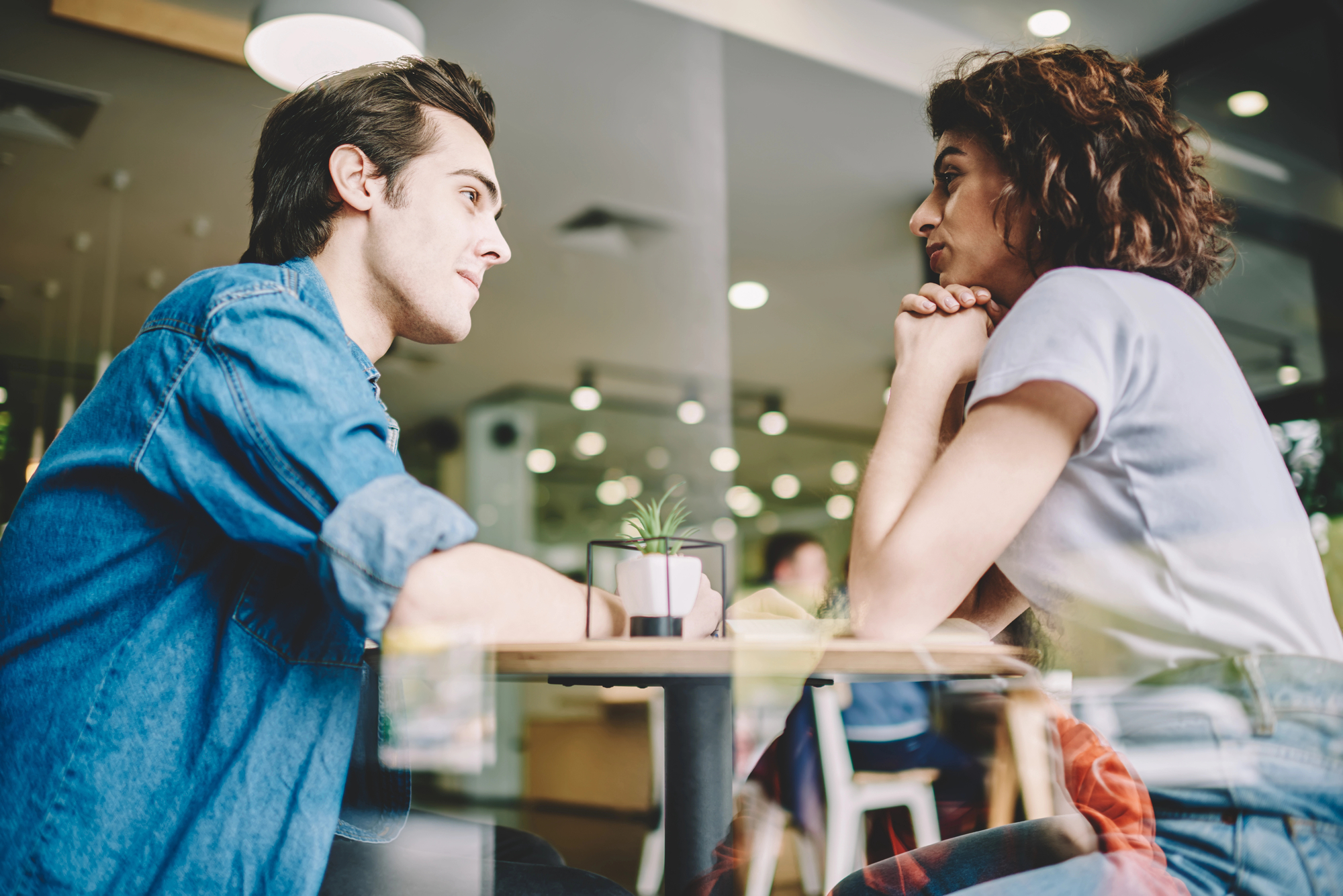 A man and a woman sit across from each other at a small table in a brightly-lit cafe. They appear to be engaged in a deep conversation, with their attention focused on each other. A small potted plant and a glass are on the table.