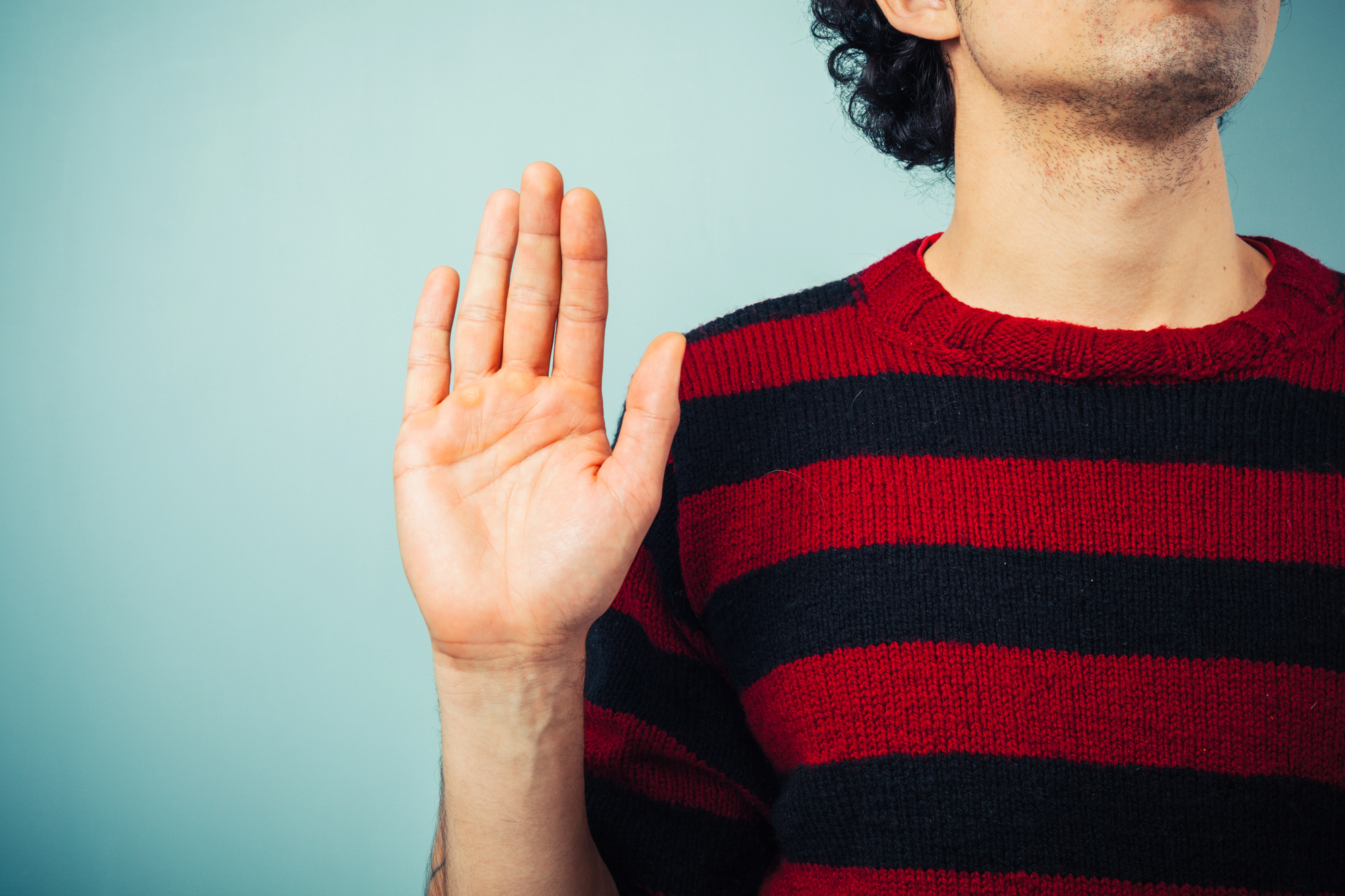 A person wearing a red and black striped sweater is shown with their right hand raised, palm facing forward, against a light blue background. The image captures the person from the shoulder up, with the person's face partially out of the frame.