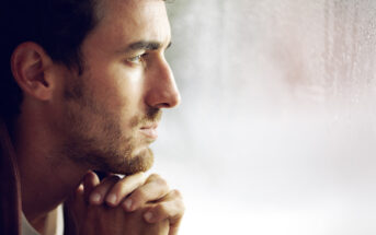 A man with short dark hair and a beard looks pensively out of a window. His hands are clasped together under his chin, and soft light illuminates his face. The window appears to have rain on it, adding to the contemplative mood.