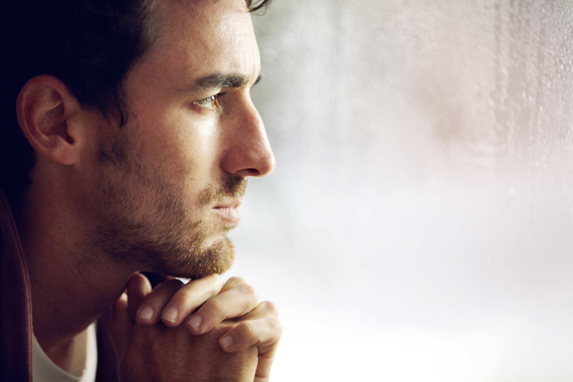 A man with short dark hair and a beard looks pensively out of a window. His hands are clasped together under his chin, and soft light illuminates his face. The window appears to have rain on it, adding to the contemplative mood.