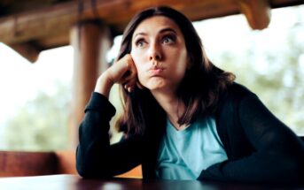 A woman with shoulder-length brown hair, wearing a blue shirt and black cardigan, is sitting at a table. She rests her head on her hand, cheeks puffed out, and gazes upward with a thoughtful or bored expression. The background is slightly blurred, indicating an outdoor setting.