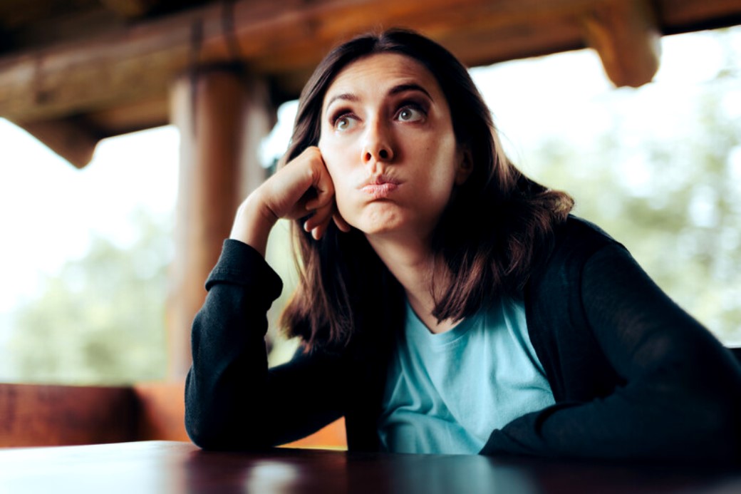 A woman with shoulder-length brown hair, wearing a blue shirt and black cardigan, is sitting at a table. She rests her head on her hand, cheeks puffed out, and gazes upward with a thoughtful or bored expression. The background is slightly blurred, indicating an outdoor setting.