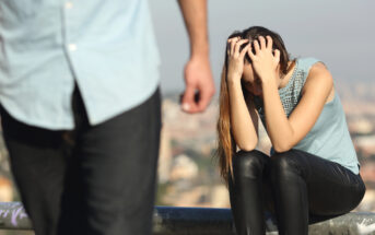 A woman sits on a rail, head in hands, appearing distressed. She is wearing a light blue sleeveless top and black pants. In the foreground, there is a person walking away, partially out of frame. The background shows a blurred cityscape.