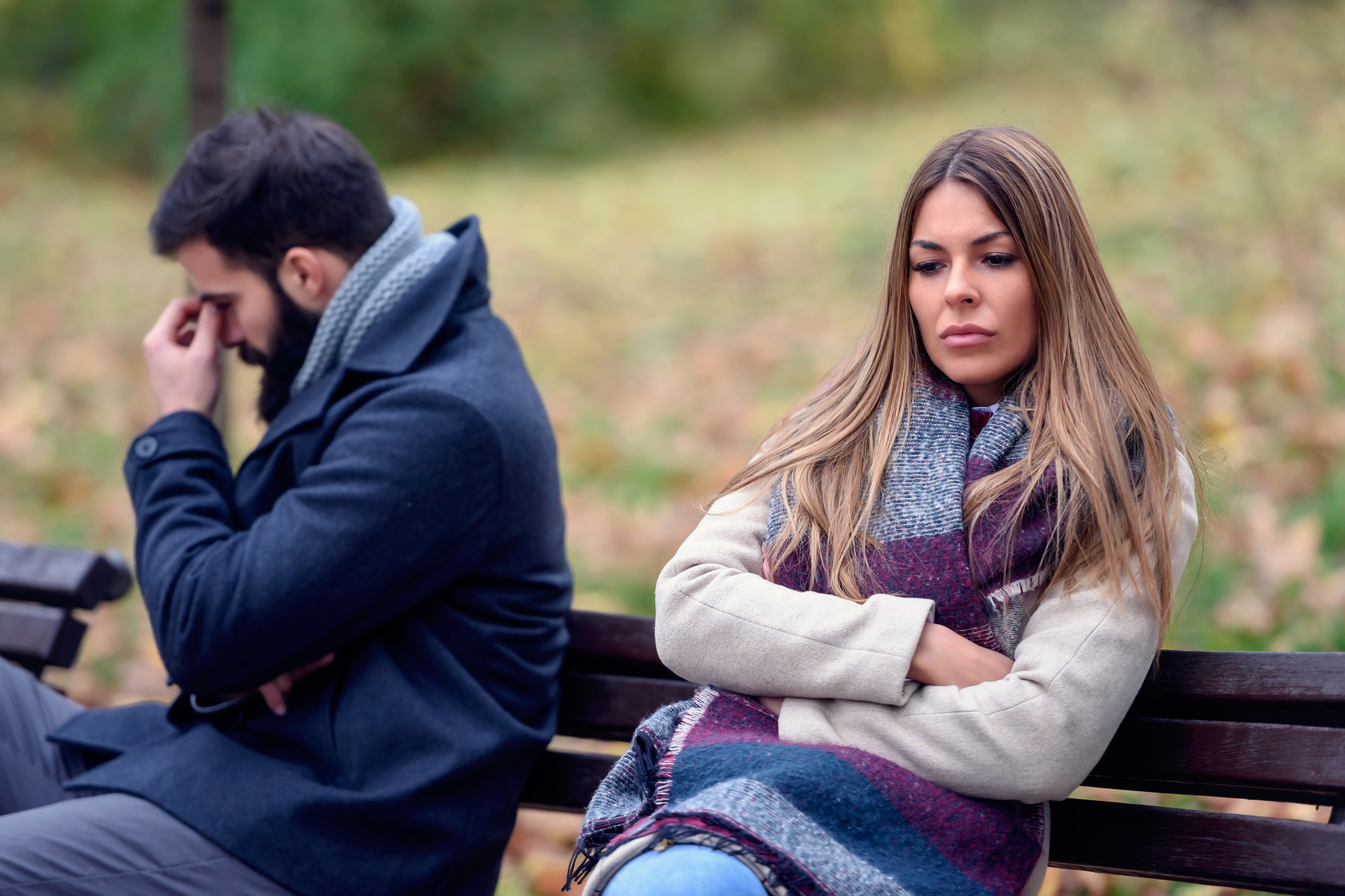 A man and a woman sit on a park bench with serious expressions. The man has his head in his hand, looking down, while the woman sits beside him with her arms crossed, staring in the opposite direction. They appear to be having a disagreement.