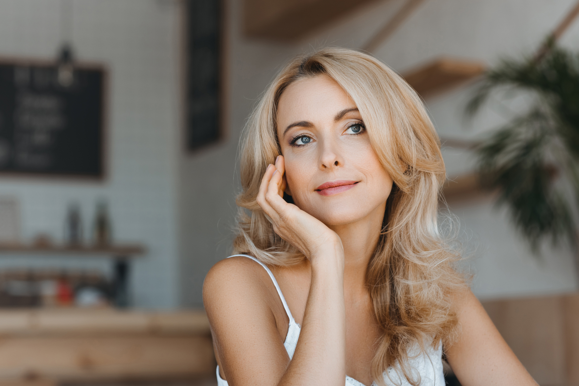 A blonde woman with wavy hair wearing a white sleeveless top, rests her chin on her hand while looking pensively into the distance. She is seated in a cozy indoor space with blurred background elements, including wooden shelves and greenery.