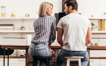 A woman and a man are sitting on stools at a kitchen counter, facing each other and having a conversation. The kitchen has a minimalist design with white walls and shelves in the background. Both individuals are wearing casual clothing.