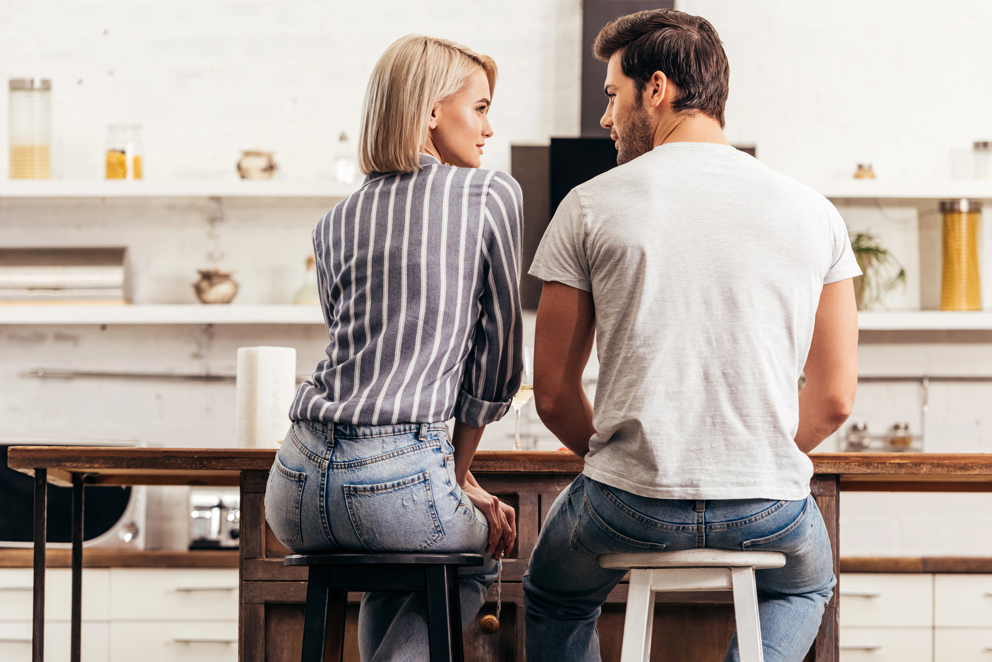 A woman and a man are sitting on stools at a kitchen counter, facing each other and having a conversation. The kitchen has a minimalist design with white walls and shelves in the background. Both individuals are wearing casual clothing.