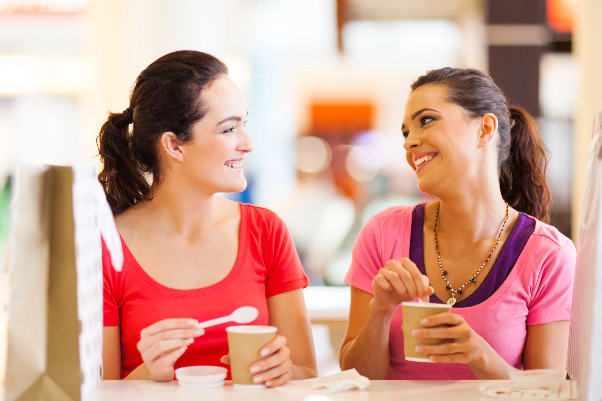 Two women sitting at a table, smiling at each other while holding cups of frozen yogurt. Both have dark hair tied back and are wearing casual t-shirts, one in red and the other in pink. Shopping bags are placed on the table, suggesting a fun day out.