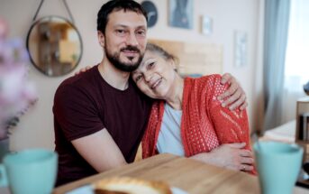 A man and an older woman, possibly family members, embrace happily while sitting at a wooden table with two green cups and a plate of food in the foreground. The setting is cozy and homey, with a round mirror and some framed pictures on the wall in the background.