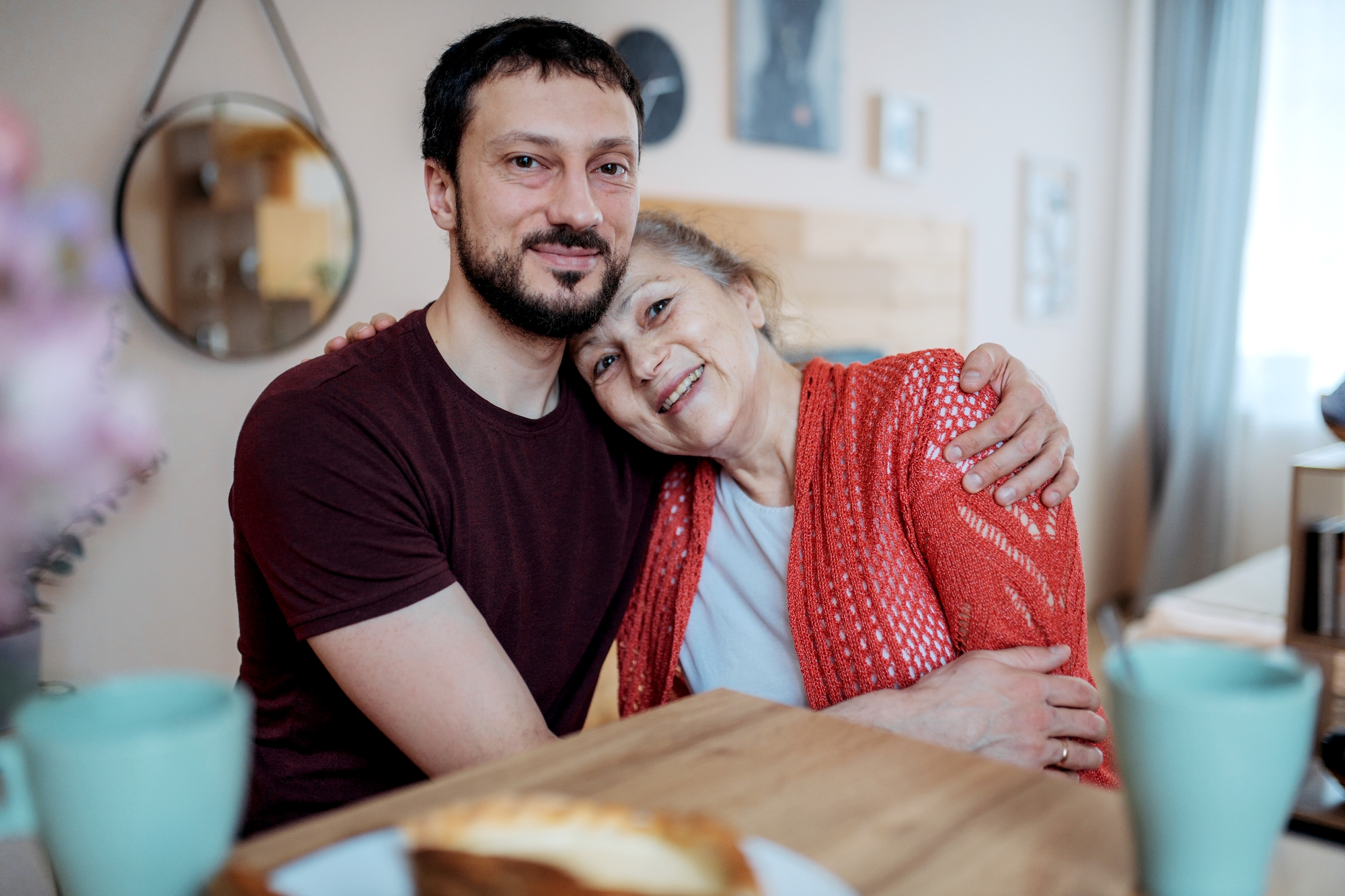 A man and an older woman, possibly family members, embrace happily while sitting at a wooden table with two green cups and a plate of food in the foreground. The setting is cozy and homey, with a round mirror and some framed pictures on the wall in the background.