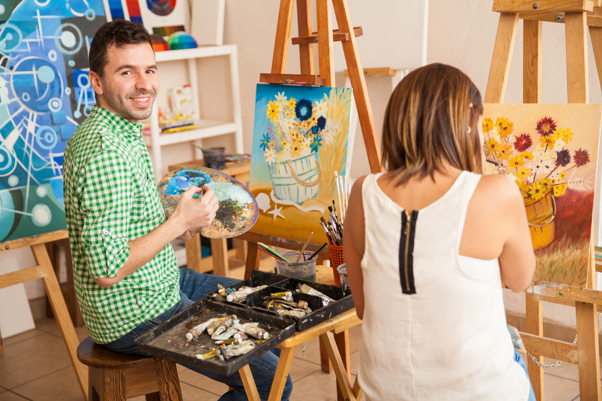 A man and a woman sitting on stools, painting on canvases mounted on easels in an art studio. The man, wearing a green checkered shirt, smiles at the camera while holding a palette and brush. The woman, in a white top, is focused on her painting of flowers.