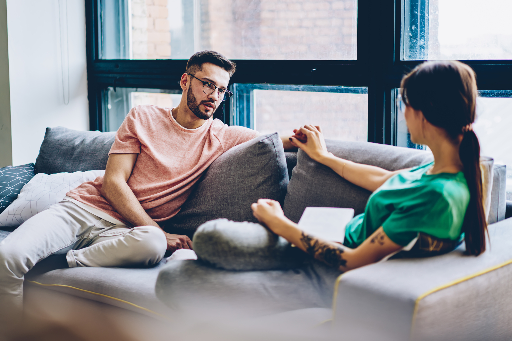 A man and a woman sit on a gray couch in a cozy living room with large windows. The man, wearing a pink shirt and glasses, is engaged in conversation with the woman, who is holding a notebook and wearing a green shirt. They appear relaxed and focused.