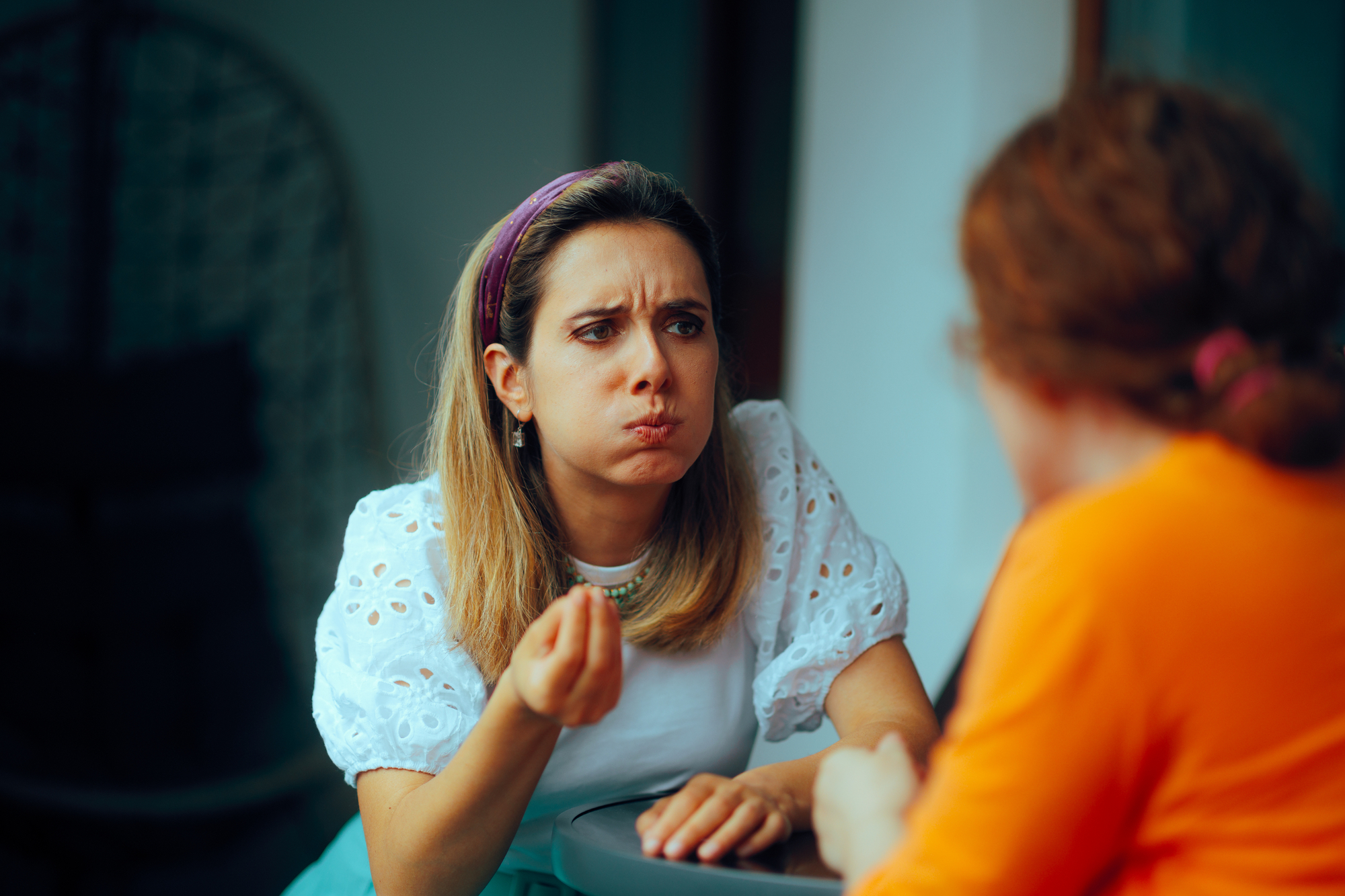 A woman with an expressive face and puffed cheeks is gesturing with her hand as she talks to another person seated across from her. She has shoulder-length hair, a headband, and is wearing a white blouse. The other person, facing away from the camera, has red hair and is wearing an orange shirt.