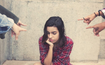 A young woman in a red plaid shirt sits against a concrete wall, looking down with a sad expression. Several hands are pointing at her from various directions, suggesting she is being accused or blamed. The scene conveys a sense of isolation and distress.
