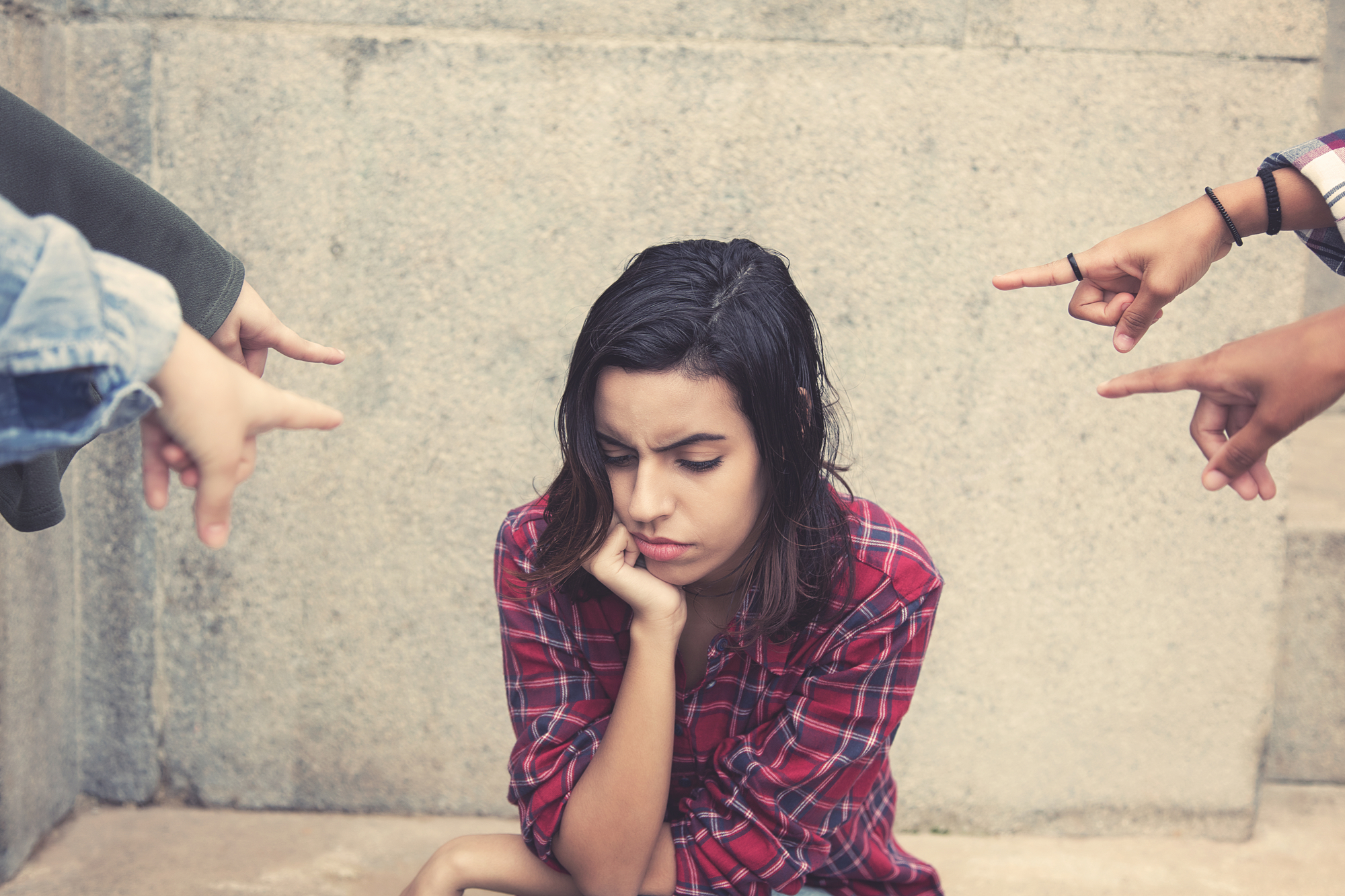 A young woman in a red plaid shirt sits against a concrete wall, looking down with a sad expression. Several hands are pointing at her from various directions, suggesting she is being accused or blamed. The scene conveys a sense of isolation and distress.