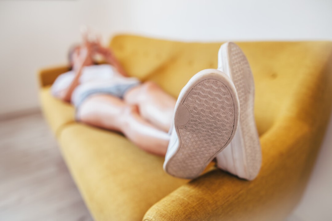 Person lying on a yellow sofa with their feet in white sneakers prominently in the foreground. The person appears to be using a smartphone, and the focus is on the shoes.